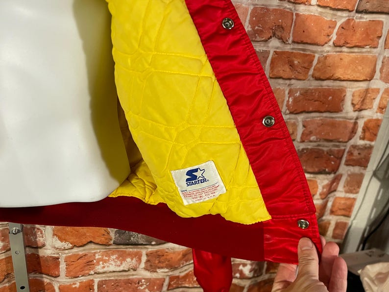 May include: Red and yellow Starter jacket with a quilted yellow lining. The jacket has a red exterior with snap closures and a red collar. The Starter logo is visible on a tag inside the jacket.