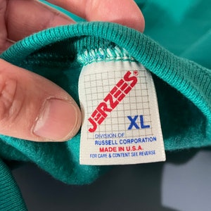 May include: A green Jerzees brand t-shirt tag with white text. The tag says "Jerzees", "Division of Russell Corporation", "Made in U.S.A.", "For care & content see reverse", and "XL".