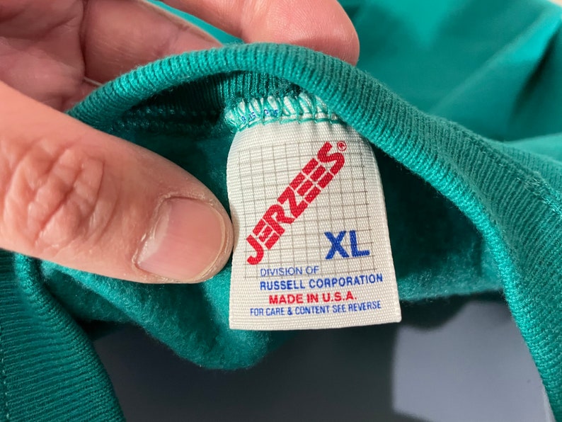 May include: A green Jerzees brand t-shirt tag with white text. The tag says "Jerzees", "Division of Russell Corporation", "Made in U.S.A.", "For care & content see reverse", and "XL".