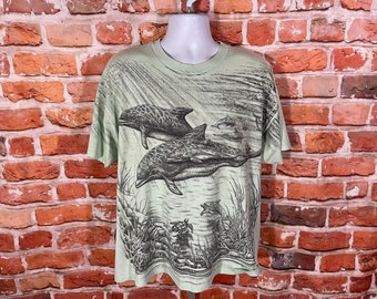 May include: A light green t-shirt with a graphic print of dolphins swimming in the ocean. The dolphins are drawn in black and white and are surrounded by seaweed and other marine life.