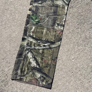 May include: Close-up of a section of camouflage pants. The fabric features a detailed pattern of browns, greens, and tans, mimicking a forest environment. The pants are lying on a concrete surface.