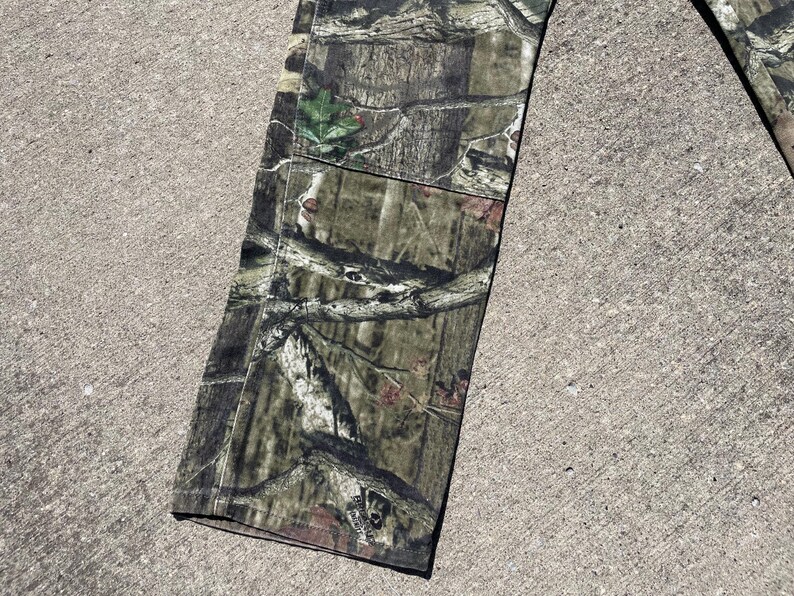 May include: Close-up of a section of camouflage pants. The fabric features a detailed pattern of browns, greens, and tans, mimicking a forest environment. The pants are lying on a concrete surface.