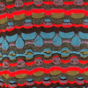 May include: Close-up of a knitted fabric with a colorful striped pattern. The fabric features a textured design with raised bumps and loops in shades of red, blue, brown, and purple.