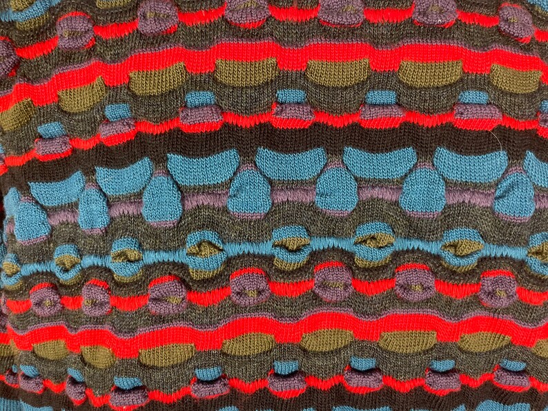 May include: Close-up of a knitted fabric with a colorful striped pattern. The fabric features a textured design with raised bumps and loops in shades of red, blue, brown, and purple.