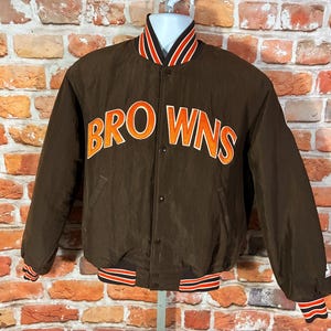 May include: A brown varsity jacket featuring the word BROWNS in orange and white lettering across the chest. The jacket has a snap-button front closure, long sleeves, and ribbed cuffs and collar with orange, white, and black striped detailing.