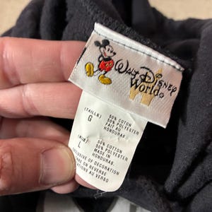 May include: Black hooded sweatshirt with a Walt Disney World tag. The tag features a Mickey Mouse graphic and the text "Walt Disney World". The tag also includes size and material information.