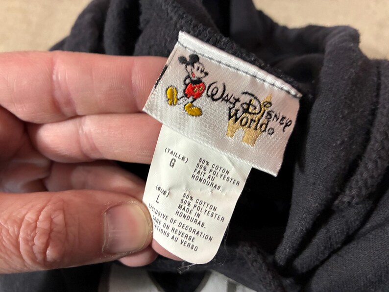 May include: Black hooded sweatshirt with a Walt Disney World tag. The tag features a Mickey Mouse graphic and the text "Walt Disney World". The tag also includes size and material information.
