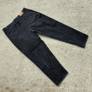 May include: A pair of black denim jeans, laid flat on a concrete surface. The jeans have a classic five-pocket design, with a faded appearance. A brown leather patch is visible on the back waistband.
