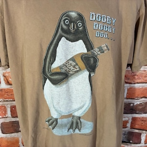 May include: A tan t-shirt featuring a graphic of a black and white penguin holding a bottle of Bud Ice beer. The text "DOOBY DOOBY DOO..." is printed in light blue above the penguin. The penguin stands on a small patch of light blue ice.