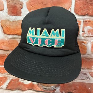 May include: A black baseball cap featuring an embroidered patch on the front panel. The patch displays the text MIAMI VICE in teal and pink lettering against a cream-colored background. The cap has a structured crown and a curved brim.