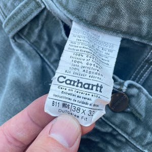 Puede incluir: Un primer plano de una etiqueta de ropa de trabajo Carhartt de ajuste dungaree. La etiqueta es verde y blanca y tiene el texto "Carhartt" en una fuente grande y en negrita. La etiqueta tambi&eacute;n incluye la talla "38 x 32" y el texto "Dungaree Fit".
