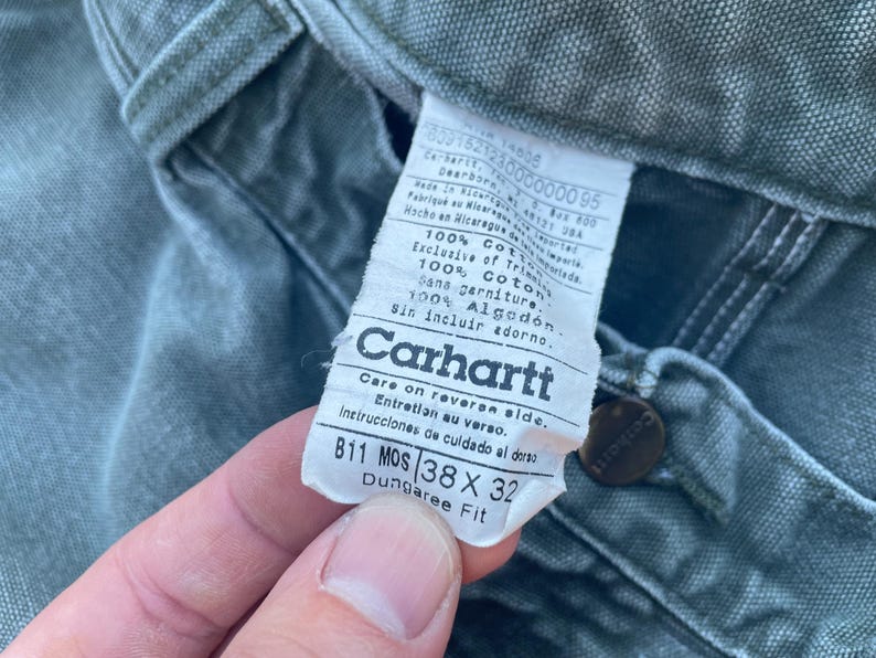 Puede incluir: Un primer plano de una etiqueta de ropa de trabajo Carhartt de ajuste dungaree. La etiqueta es verde y blanca y tiene el texto "Carhartt" en una fuente grande y en negrita. La etiqueta tambi&eacute;n incluye la talla "38 x 32" y el texto "Dungaree Fit".
