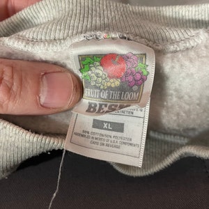 Puede incluir: Una sudadera gris Fruit of the Loom con una etiqueta blanca. La etiqueta dice "Fruit of the Loom" y "Best" con un logotipo de una manzana roja y uvas. La etiqueta tambi&eacute;n dice "XL", "50% Algod&oacute;n/50% Poli&eacute;ster", "Ensamblado en M&eacute;xico con componentes de EE. UU." y "Cuidado en el reverso."