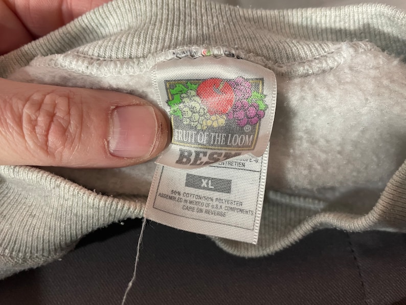 Puede incluir: Una sudadera gris Fruit of the Loom con una etiqueta blanca. La etiqueta dice "Fruit of the Loom" y "Best" con un logotipo de una manzana roja y uvas. La etiqueta tambi&eacute;n dice "XL", "50% Algod&oacute;n/50% Poli&eacute;ster", "Ensamblado en M&eacute;xico con componentes de EE. UU." y "Cuidado en el reverso."