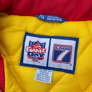 May include: A quilted jacket interior featuring a bright yellow lining and a red collar. A blue and white label displays the NFL Game Day and Logo 7 branding. A smaller white tag below indicates size XL/XG/TG, RN#63388, CA#13924, and Made in Korea.