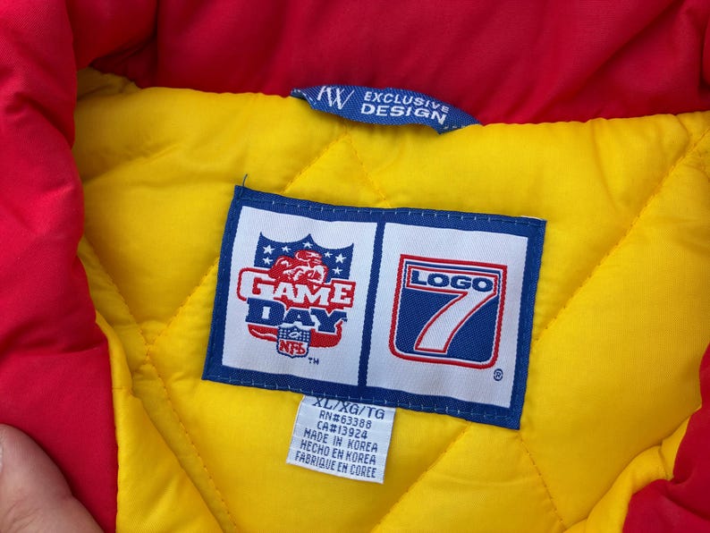 May include: A quilted jacket interior featuring a bright yellow lining and a red collar. A blue and white label displays the NFL Game Day and Logo 7 branding. A smaller white tag below indicates size XL/XG/TG, RN#63388, CA#13924, and Made in Korea.
