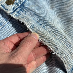 Peut inclure: Gros plan sur un tissu denim bleu clair avec des signes d'usure visibles. L'image met en &eacute;vidence la texture du denim, y compris les coutures et les bords effiloch&eacute;s. Une main touche le tissu, soulignant les d&eacute;tails du mat&eacute;riau.