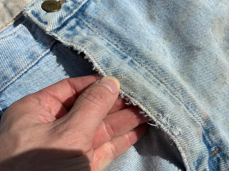 Peut inclure: Gros plan sur un tissu denim bleu clair avec des signes d'usure visibles. L'image met en &eacute;vidence la texture du denim, y compris les coutures et les bords effiloch&eacute;s. Une main touche le tissu, soulignant les d&eacute;tails du mat&eacute;riau.