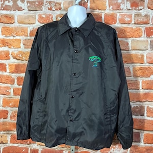 May include: Black nylon coach jacket with a button-up front, collar, and long sleeves. The jacket features a green and blue graphic on the left chest. The jacket is hanging on a white mannequin head against a brick wall.