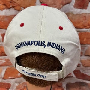 Op de afbeelding: Een beige honkbalpet met de geborduurde tekst Indianapolis, Indiana in donkerblauwe letters. De verstelbare achterband bevat de geborduurde tekst Members Only in donkerblauw. De pet heeft rode oogjes en een rode knoop aan de bovenkant.