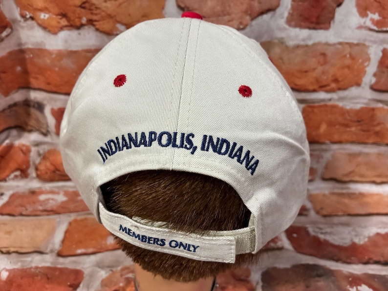 Op de afbeelding: Een beige honkbalpet met de geborduurde tekst Indianapolis, Indiana in donkerblauwe letters. De verstelbare achterband bevat de geborduurde tekst Members Only in donkerblauw. De pet heeft rode oogjes en een rode knoop aan de bovenkant.