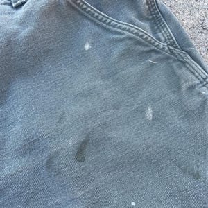 Puede incluir: Primer plano de un par de pantalones de trabajo grises con varias salpicaduras de pintura blanca.