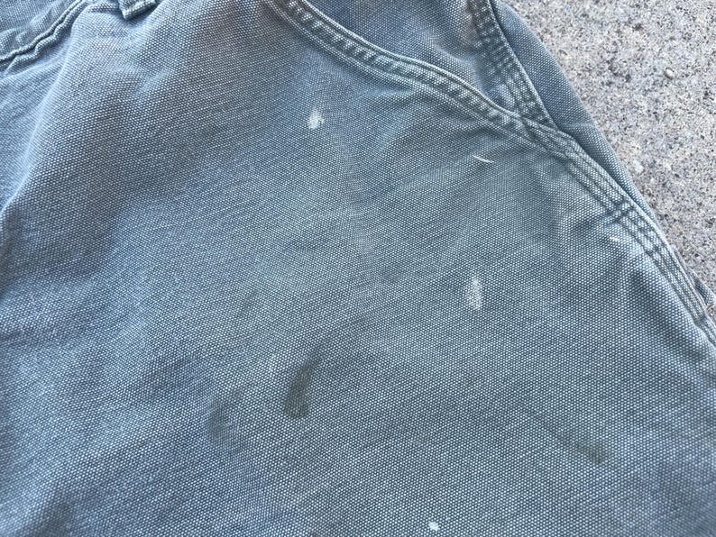 Puede incluir: Primer plano de un par de pantalones de trabajo grises con varias salpicaduras de pintura blanca.