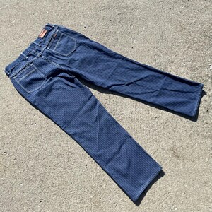 May include: Dark blue denim jeans with vertical striped pattern. The jeans have white stitching, back pockets, and a small red tag. The jeans are laid out on a concrete surface.