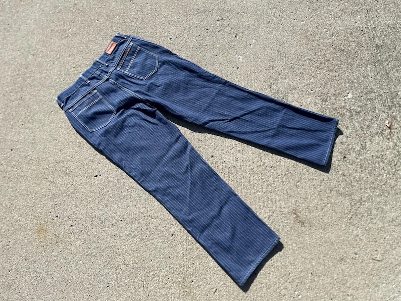 May include: Dark blue denim jeans with vertical striped pattern. The jeans have white stitching, back pockets, and a small red tag. The jeans are laid out on a concrete surface.