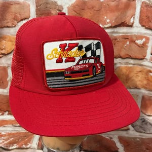 May include: A red trucker hat with a mesh back and a curved brim. The front features a rectangular patch with the name "Schader" in gold lettering, a race car, and the number "25".