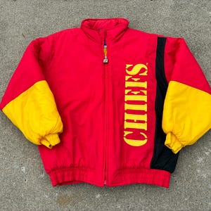 May include: A red, yellow, and black puffer jacket featuring a high collar, a full-length front zipper, and long sleeves with yellow sections. The word CHIEFS is printed vertically in large yellow capital letters on the right side of the chest.