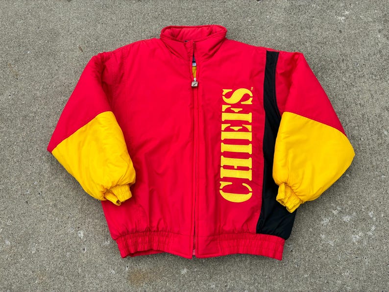 May include: A red, yellow, and black puffer jacket featuring a high collar, a full-length front zipper, and long sleeves with yellow sections. The word CHIEFS is printed vertically in large yellow capital letters on the right side of the chest.