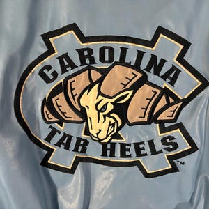 May include: Light blue jacket with a large embroidered logo. The logo features a ram's head in tan and black, surrounded by the words "CAROLINA TAR HEELS" in black and white.
