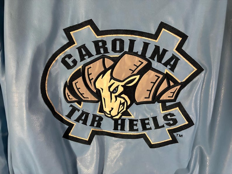 May include: Light blue jacket with a large embroidered logo. The logo features a ram's head in tan and black, surrounded by the words "CAROLINA TAR HEELS" in black and white.