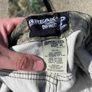 May include: Close-up of camouflage pants with a tag that reads "BREAK UP INFINITY" and "MOSSY OAK". The tag also indicates the size is 36/34 and the pants are made of 100% cotton. The UPC is #4273304603.