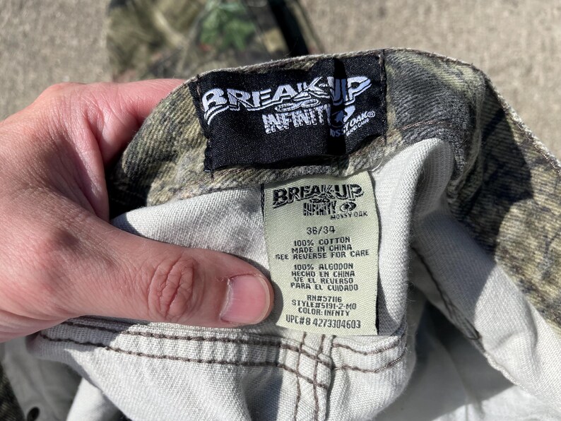 May include: Close-up of camouflage pants with a tag that reads "BREAK UP INFINITY" and "MOSSY OAK". The tag also indicates the size is 36/34 and the pants are made of 100% cotton. The UPC is #4273304603.