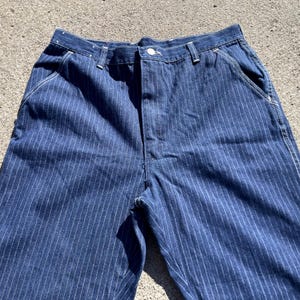 May include: Close-up of blue denim pants with vertical white pinstripes. The pants have a button closure, belt loops, and front pockets. The fabric appears to be a medium-weight denim, suitable for casual wear. The pants are unhemmed.