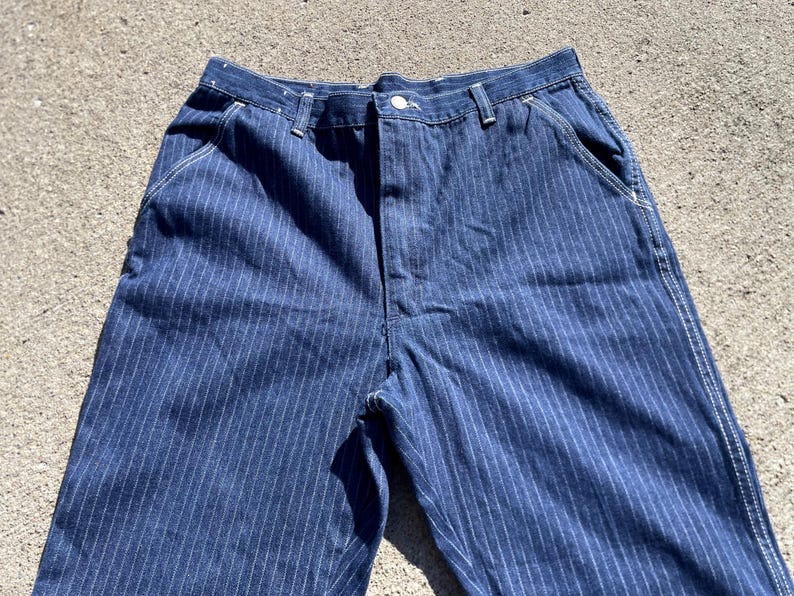 May include: Close-up of blue denim pants with vertical white pinstripes. The pants have a button closure, belt loops, and front pockets. The fabric appears to be a medium-weight denim, suitable for casual wear. The pants are unhemmed.