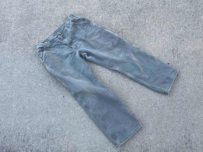 Puede incluir: Un par de pantalones de trabajo grises con un aspecto descolorido. Los pantalones tienen una pierna recta y est&aacute;n hechos de una tela duradera.