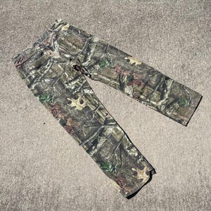 May include: Camouflage-patterned pants in shades of green, brown, and tan. The pants are made of a durable fabric and feature a classic five-pocket design. The camouflage pattern is designed to blend in with a wooded environment.