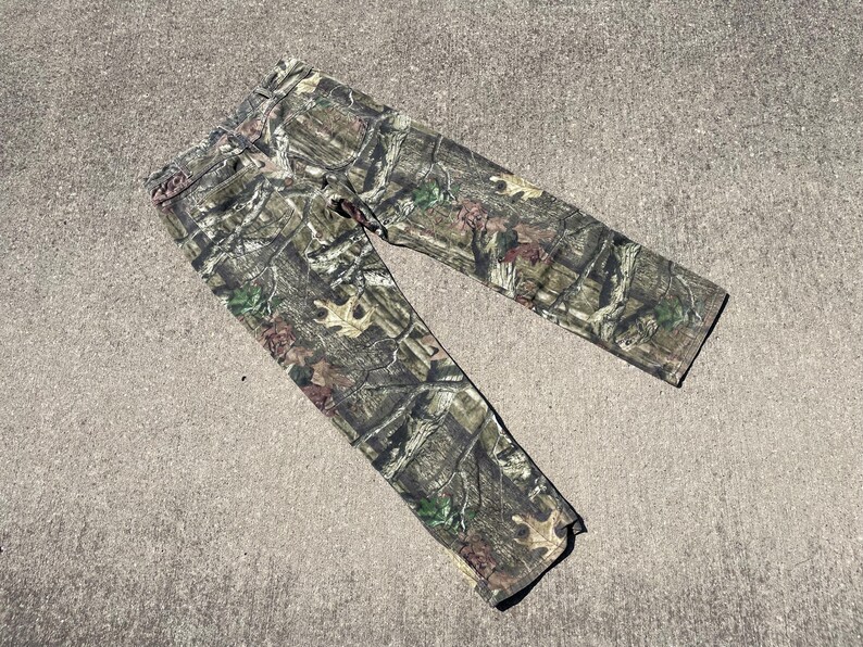 May include: Camouflage-patterned pants in shades of green, brown, and tan. The pants are made of a durable fabric and feature a classic five-pocket design. The camouflage pattern is designed to blend in with a wooded environment.