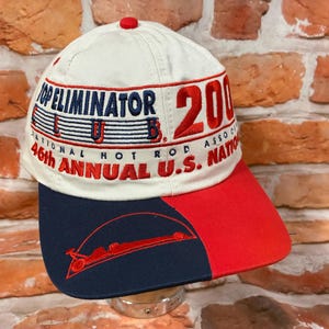 Op de afbeelding: Een cr&egrave;mekleurige baseballpet met geborduurde tekst die Top Eliminator Club 2000 en 46th Annual U.S. Nationals leest. De pet heeft een tweekleurige klep in marineblauw en rood, met een rode afbeelding van een dragster op het marineblauwe deel.