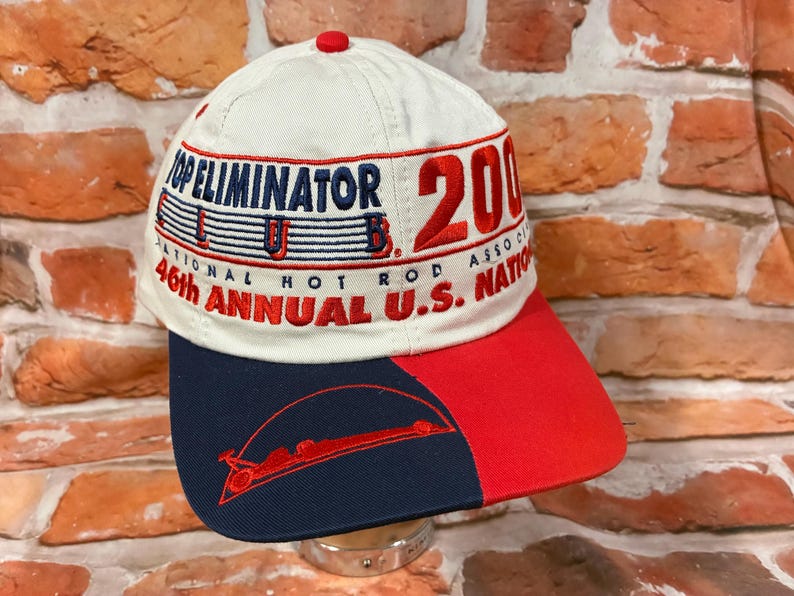 Op de afbeelding: Een cr&egrave;mekleurige baseballpet met geborduurde tekst die Top Eliminator Club 2000 en 46th Annual U.S. Nationals leest. De pet heeft een tweekleurige klep in marineblauw en rood, met een rode afbeelding van een dragster op het marineblauwe deel.