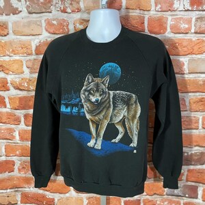 Op de afbeelding: Zwart sweatshirt met een gedetailleerde illustratie van een wolf onder een sterrenhemel en een blauwe maan. De wolf is afgebeeld in bruin en grijs, staand op een blauw pad. Het ontwerp bevat een bos silhouet.