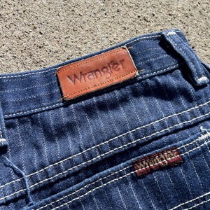 May include: Close-up of blue denim jeans with white pinstripes and white stitching. The brown leather Wrangler label is visible at the waistband. The Wrangler logo is also on the back pocket.