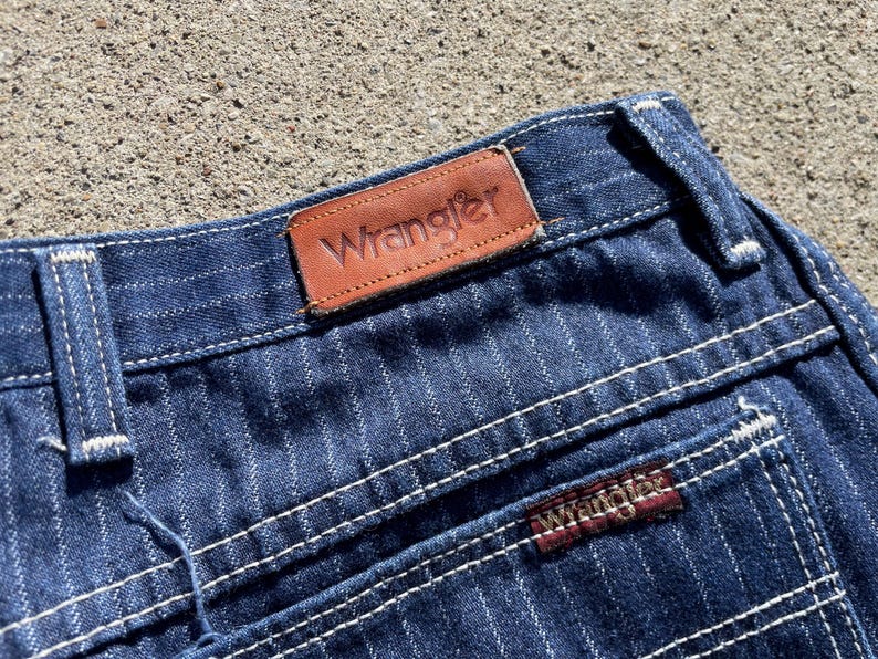 May include: Close-up of blue denim jeans with white pinstripes and white stitching. The brown leather Wrangler label is visible at the waistband. The Wrangler logo is also on the back pocket.