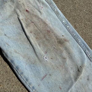 Peut inclure: Gros plan sur un jean en denim bleu clair, montrant des signes d'usure. Le jean pr&eacute;sente des taches et des &eacute;claboussures de peinture de diff&eacute;rentes couleurs, dont le rouge et le gris. Les coutures sont visibles.