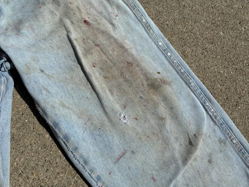 Peut inclure: Gros plan sur un jean en denim bleu clair, montrant des signes d'usure. Le jean pr&eacute;sente des taches et des &eacute;claboussures de peinture de diff&eacute;rentes couleurs, dont le rouge et le gris. Les coutures sont visibles.