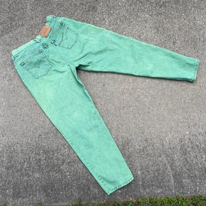 May include: Mint green denim jeans with a classic five-pocket design. The jeans feature a faded wash and a brown leather patch on the back. The jeans are laid out on a concrete surface.