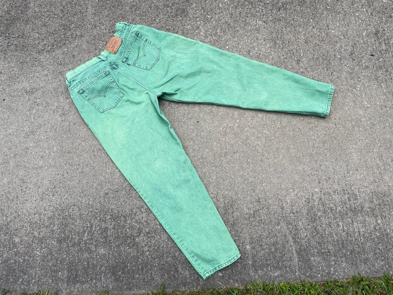 May include: Mint green denim jeans with a classic five-pocket design. The jeans feature a faded wash and a brown leather patch on the back. The jeans are laid out on a concrete surface.